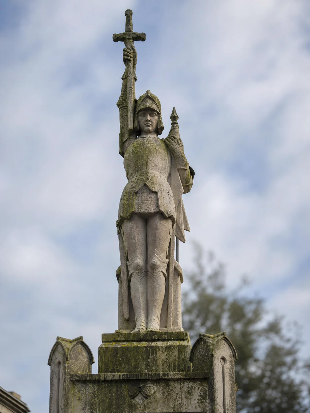 Jeanne d'Arc en armure, l'épée levée et l'étendard à la main. La pierre a pris le lichen ; le bras droit n'est pas d'origine.